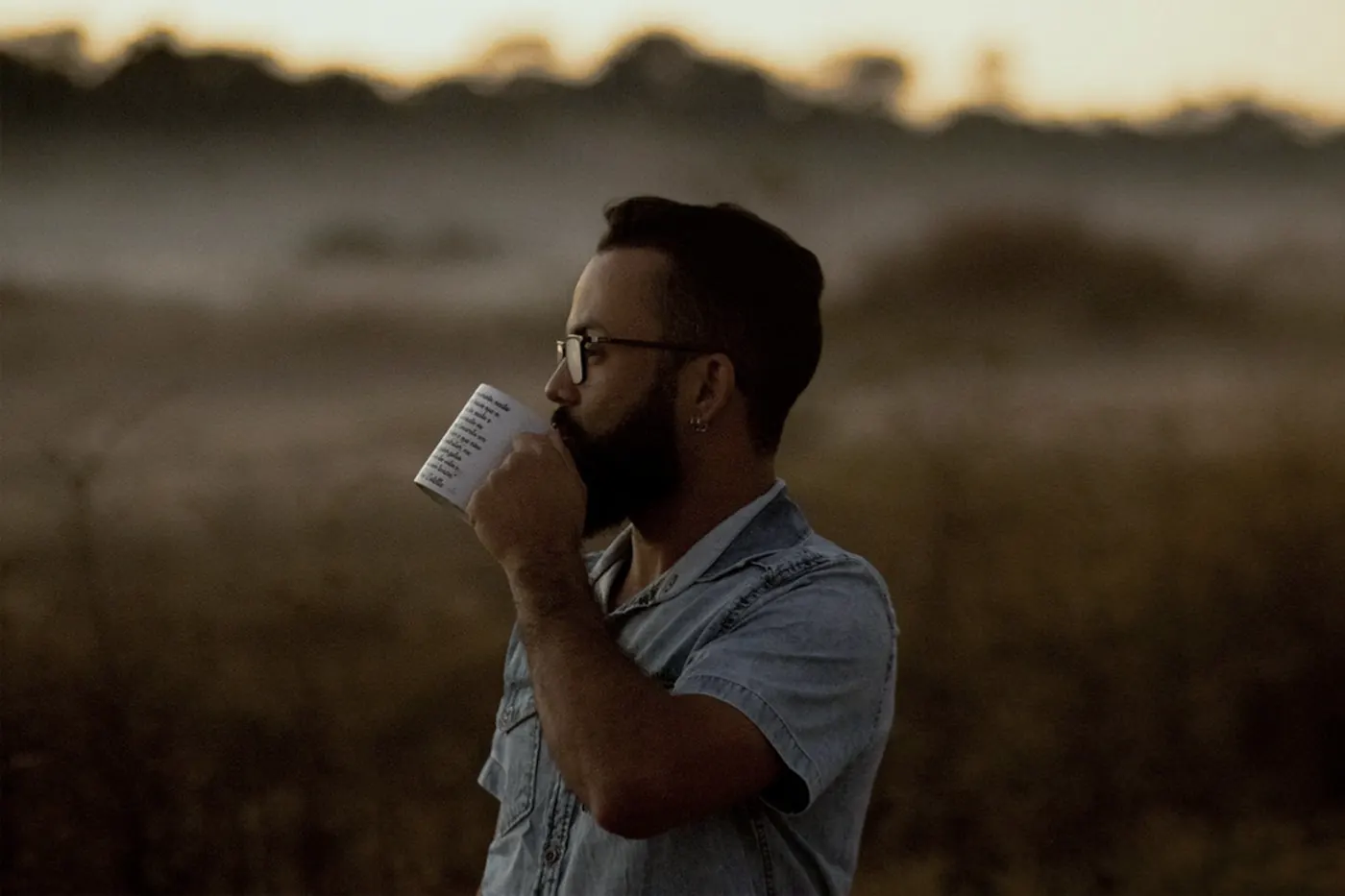 A bearded man drinking coffee outdoors in a field at sunrise