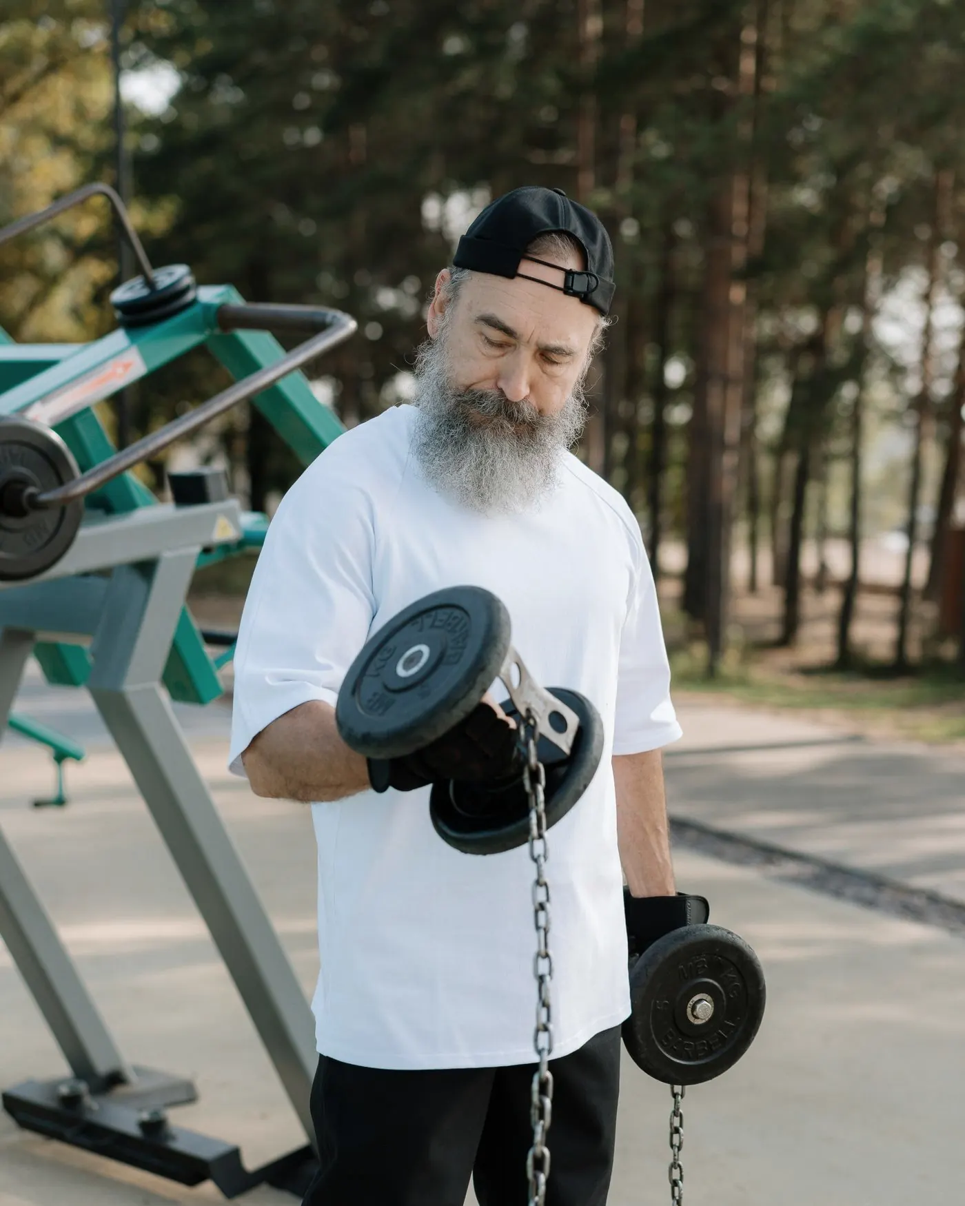 A bearded man in a white T-shirt doing a dumbbell curl at an outdoor gym among pine trees